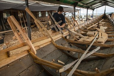 Bådebyggerlærling Magnus Ibsen Rishøj monterer en af de lange bjælkevægere. Under arbejdet holdes vægeren på plads med bordklemmer, mens placering og tilpasning kontrolleres langs skibets fulde længde, inden vægeren fastgøres med trænagler. Bådebyggerlærling Magnus Ibsen Rishøj monterer en af de lange bjælkevægere. Under arbejdet holdes vægeren på plads med bordklemmer, mens placering og tilpasning kontrolleres langs skibets fulde længde, inden vægeren fastgøres med trænagler.